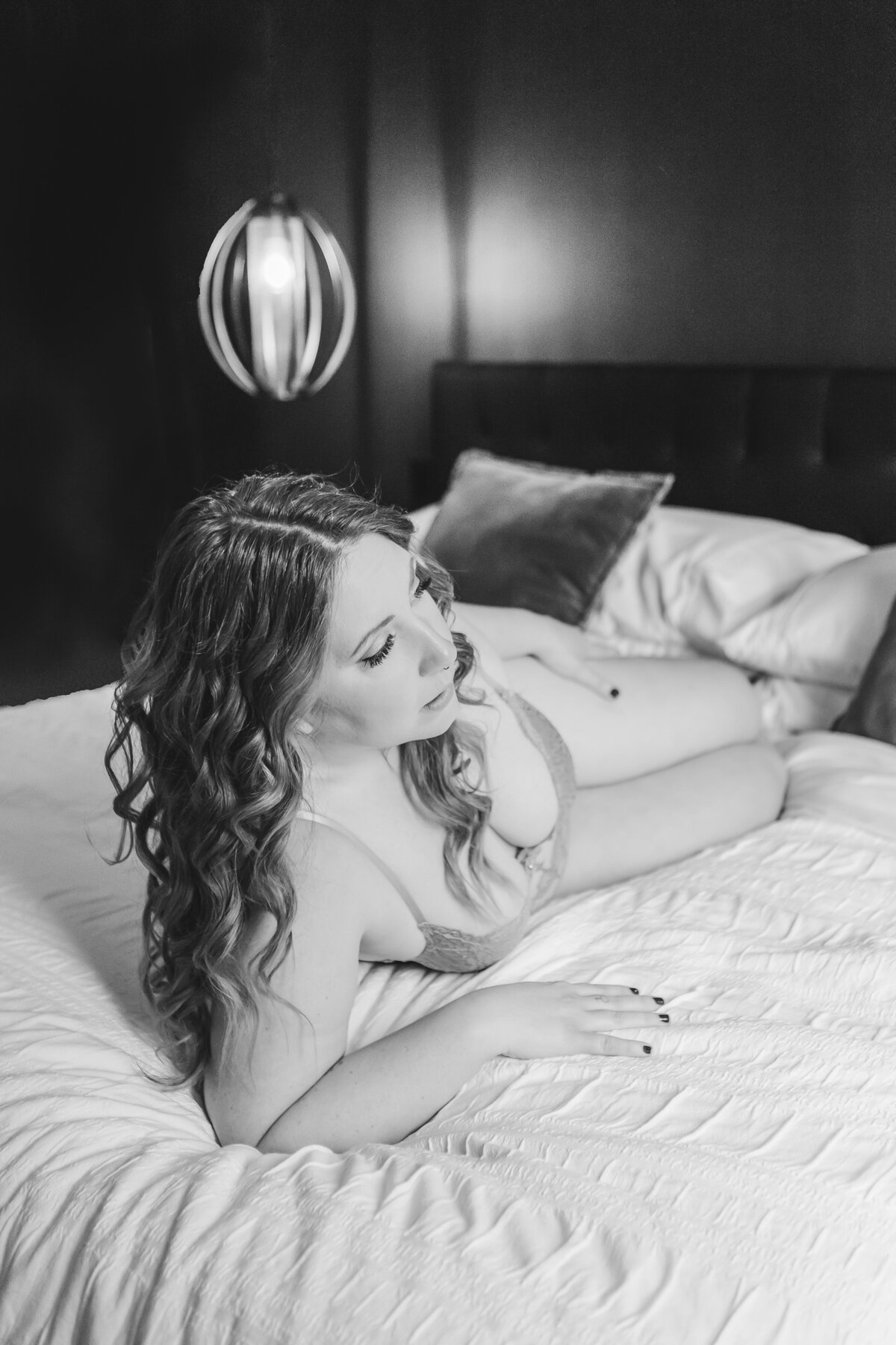 A woman lying on a bed in light gray lingerie, lit by warm lights, looking dreamily off into the distance.