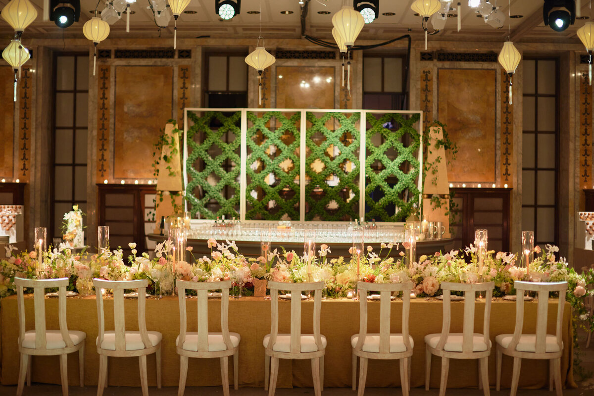This beautifully decorated reception hall features an elegantly set long dining table with floral arrangements and candles. Behind the table, a decorative green wall with foliage adds to the ambiance. Soft lighting and hanging lanterns create a warm, inviting atmosphere. This stunning scene was captured at the New York Public Library in New York City by photographer Shawn Connell, and videographers Aaron Navak Films.