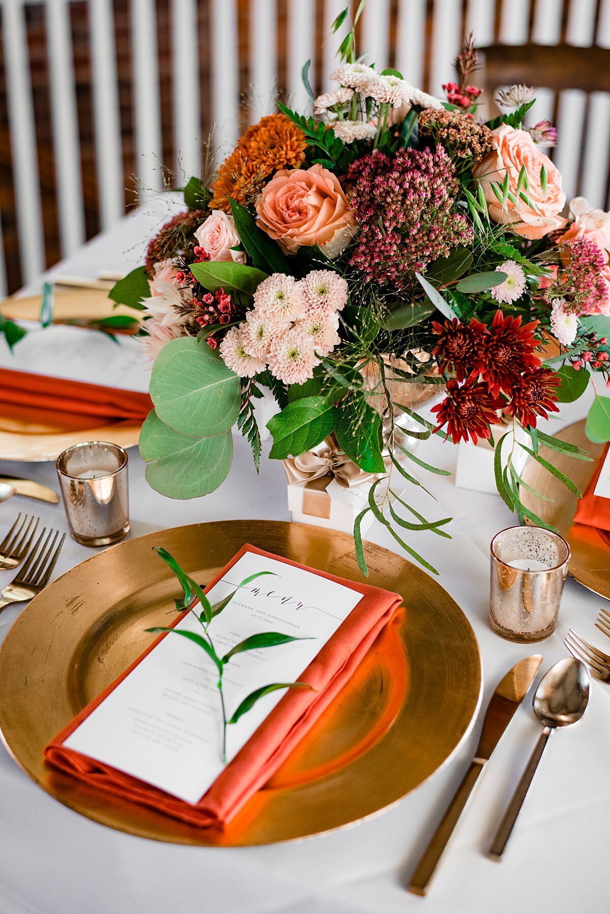 A table covered with a white linen is set with a gold charger and a folded rust colored napkin with a menu card and sprig of greenery. On either side of the plate is golf flatware and gold votive candles. At the center of the table is a large round centerpiece of eucalyptus, peach roses, burgundy mums, blush mums, blush tea roses, orange mums and ivory anemone.