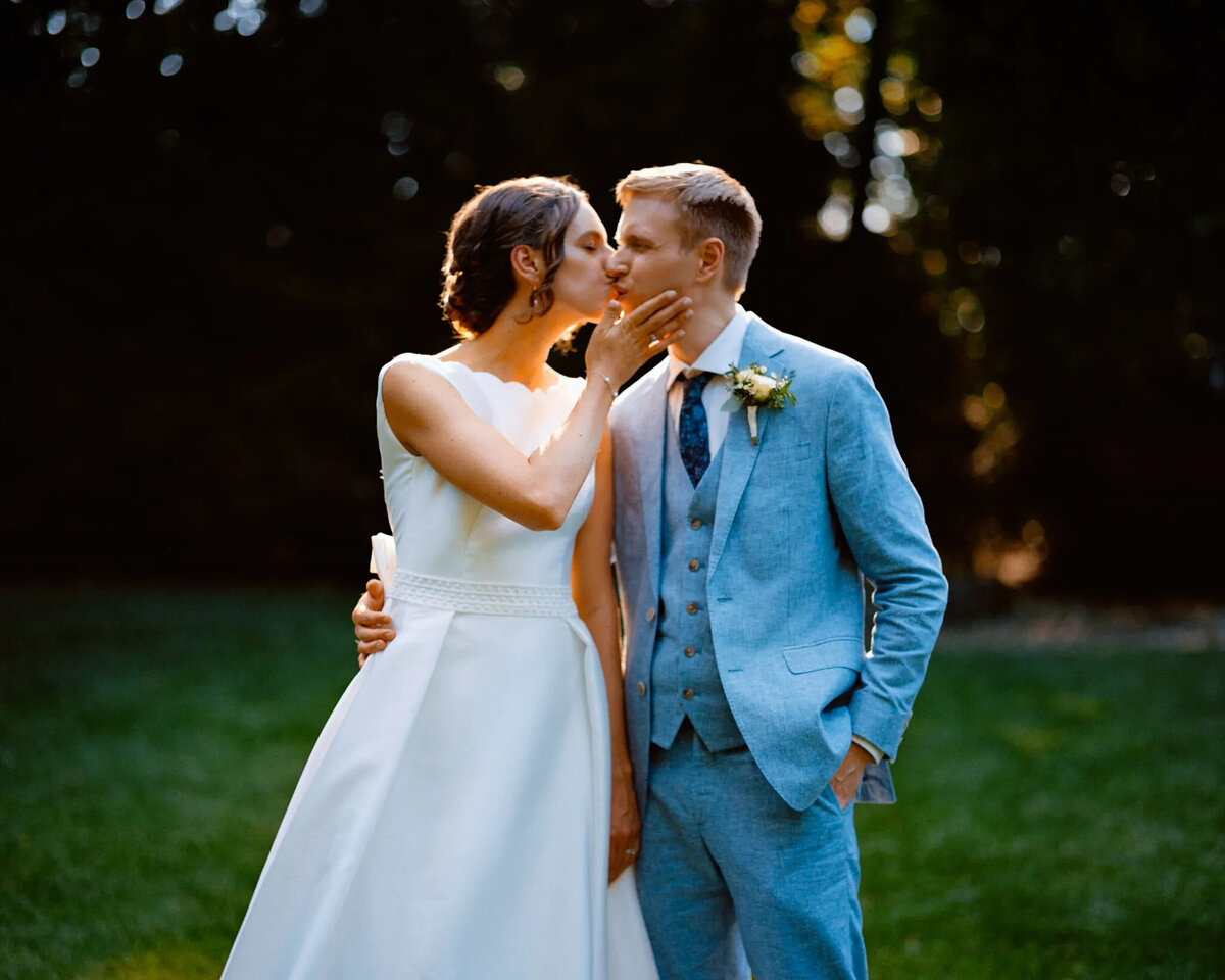 A bride in a white dress gently kisses the forehead of a groom in a light blue suit as they stand together outdoors on a grassy lawn, surrounded by trees and soft sunlight, beautifully captured by an NJ wedding photographer.