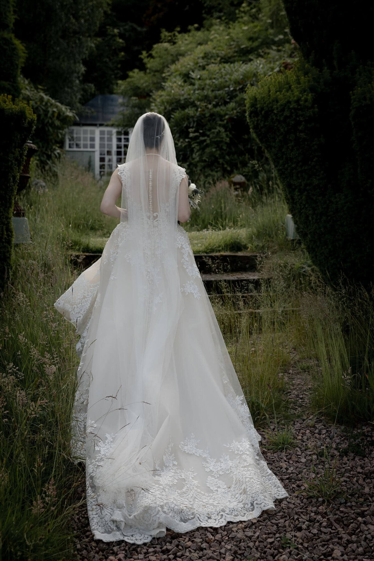 A bride walks through a pathway of large bushes up some steps.