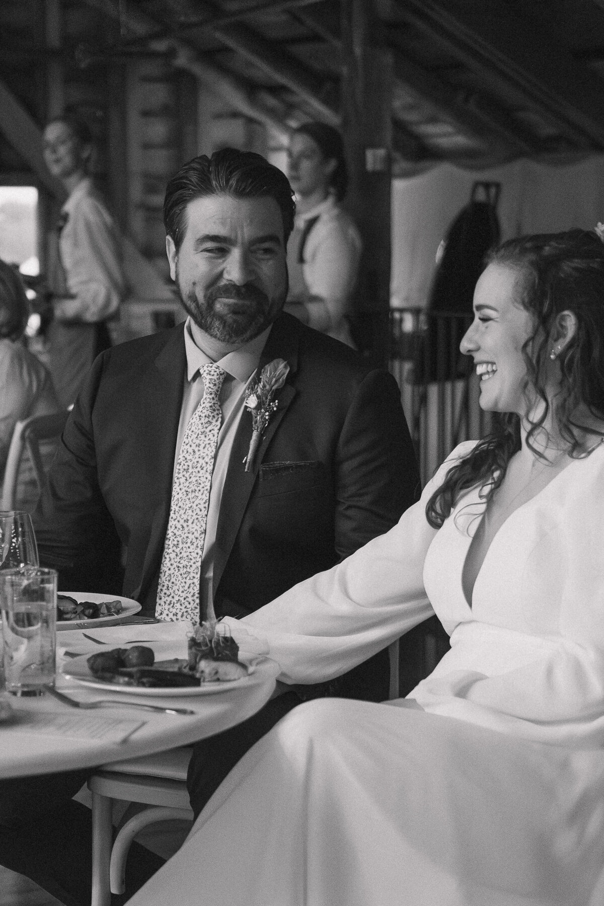 Bride and groom share a joyful moment at their wedding reception, captured in black and white. The bride, wearing a long-sleeved gown, and the groom, in a suit with a floral tie, laugh together as they enjoy their meal. This candid, documentary-style photo highlights their happiness and the intimate atmosphere of the celebration.