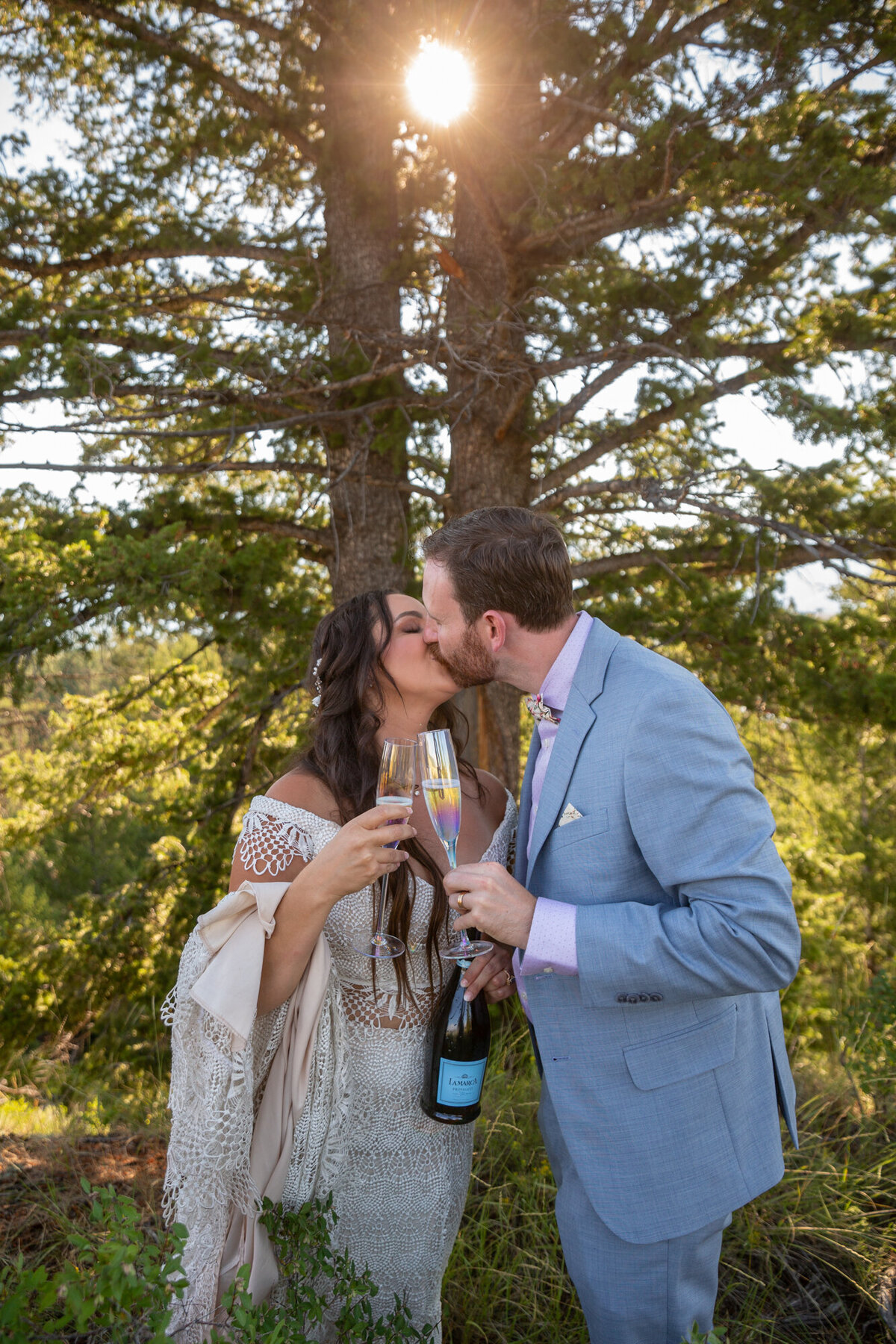 A couple holding a champagne bottle kiss as the sun flares through the trees behind them.