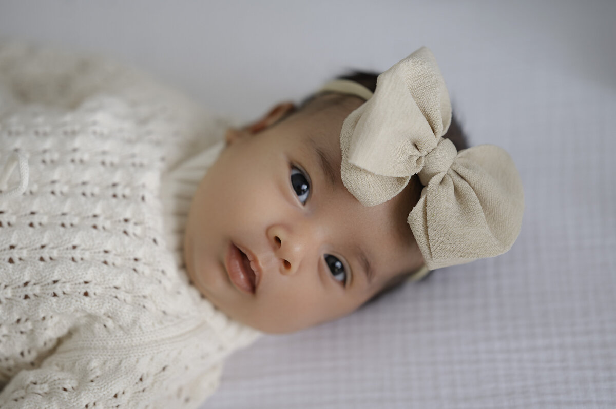 Newborn baby girl with a beige bow looks right into the camera with alert eyes