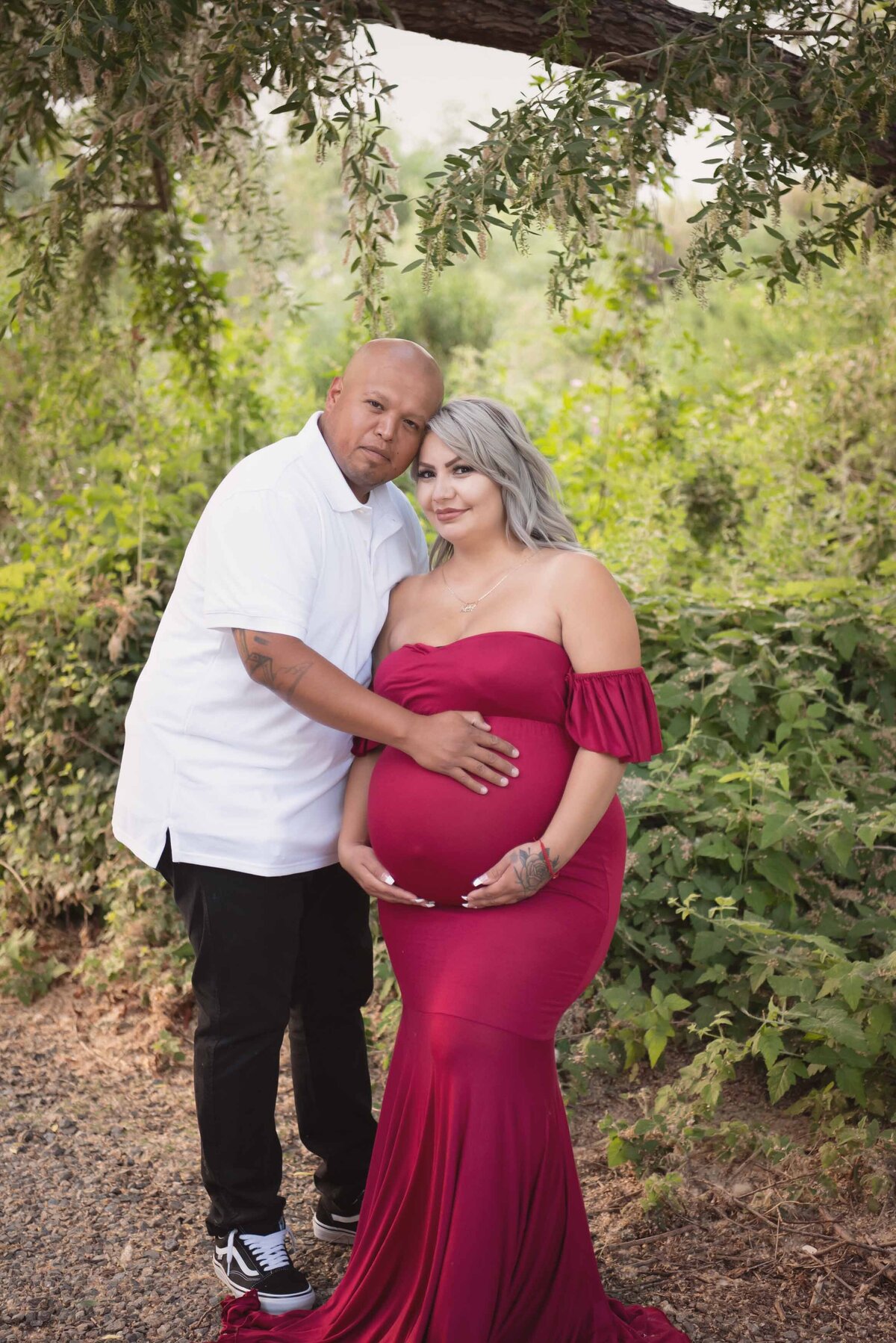 Expectant Mom and Dad in park smiling at camera and holding their baby bump. Mom is wearing a long, red maternity gown.