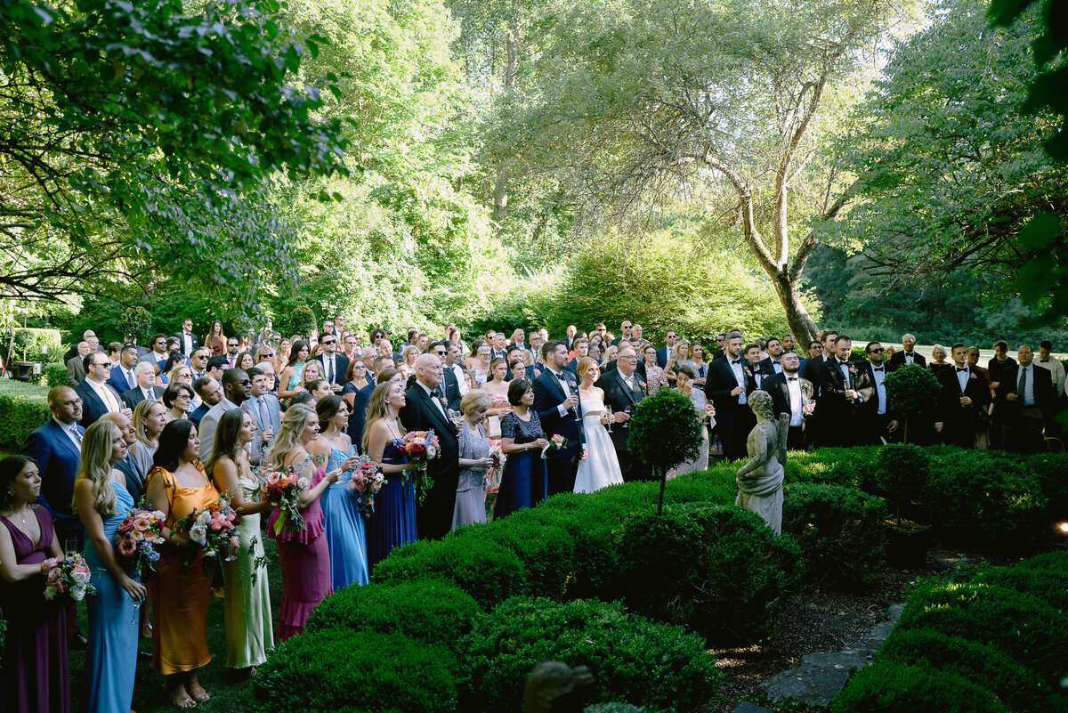 Guests gather holding drinks amongst greenery for toasts during cocktail hour at the Lord Thompson Manor
