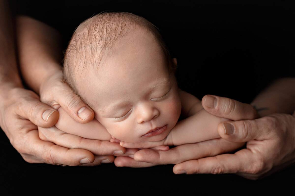 newborn baby laying in mom and dads hands on a black background