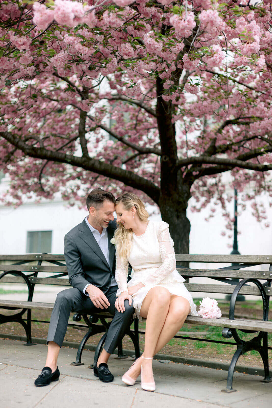 The eloping bride and groom, are happily sitting on a bench in NY Central Park with their heads touching. Image by Jenny Fu Studio