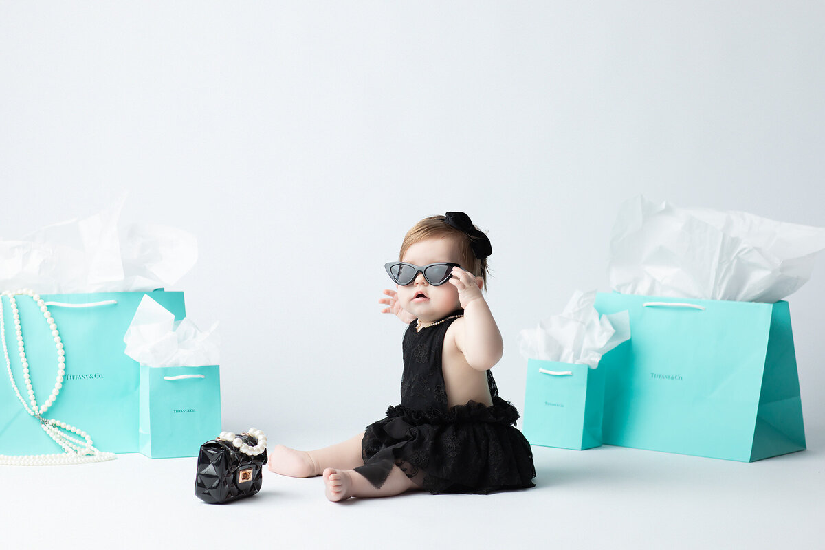 Dallas milestone photographer features a baby girl dressed like Audrey Hepburn with cat-eye sunglasses, pearls, and a black lace outfit, sitting among Tiffany blue gift bags and a toy purse.