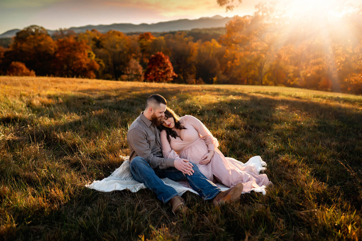 An expecting couple has a quiet, snuggly moment on a blanket in the grass as the sun sets behind them