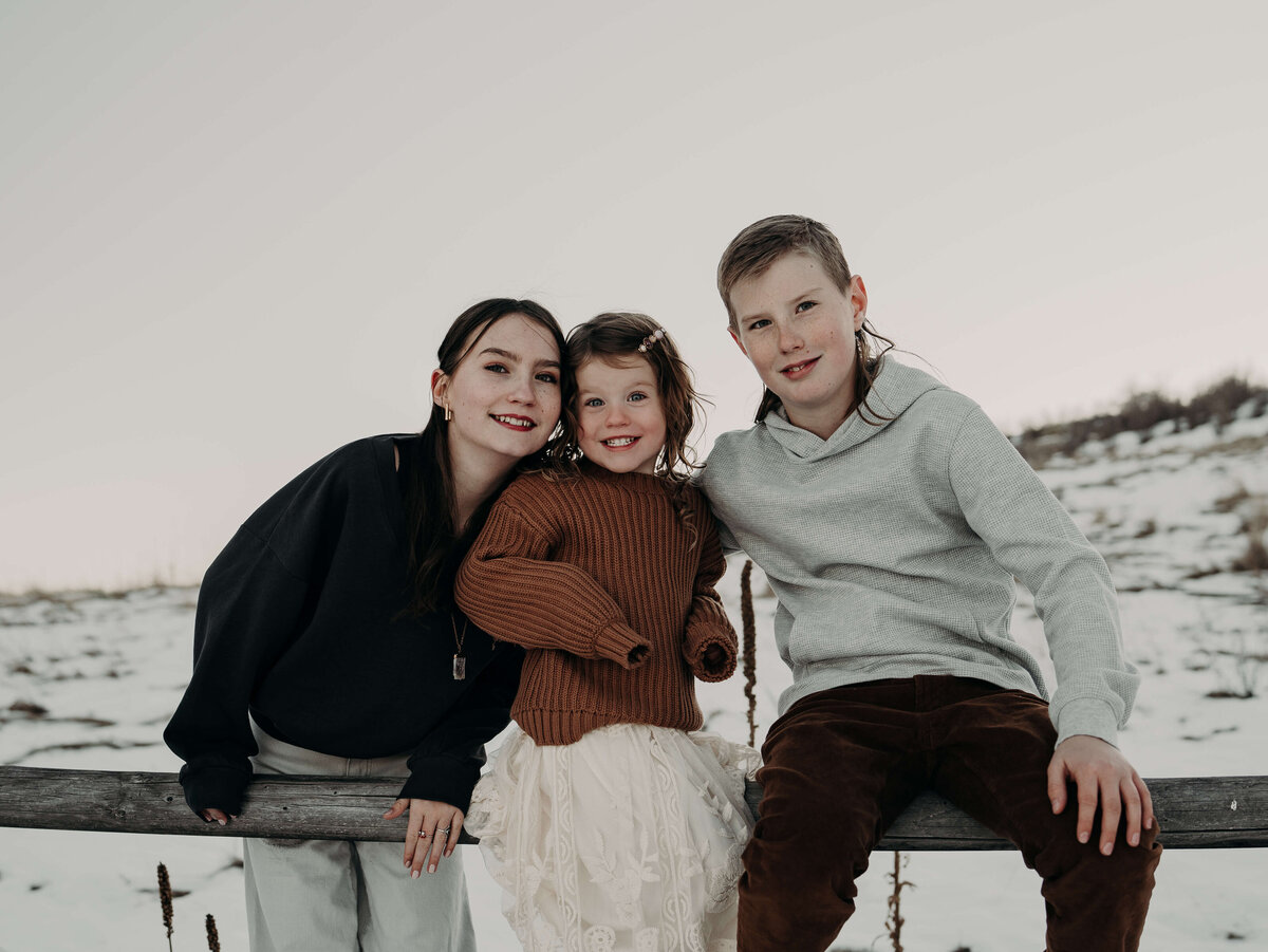 Three siblings sit on a fence on a family farm, wearing perfectly coordinated neutral tones