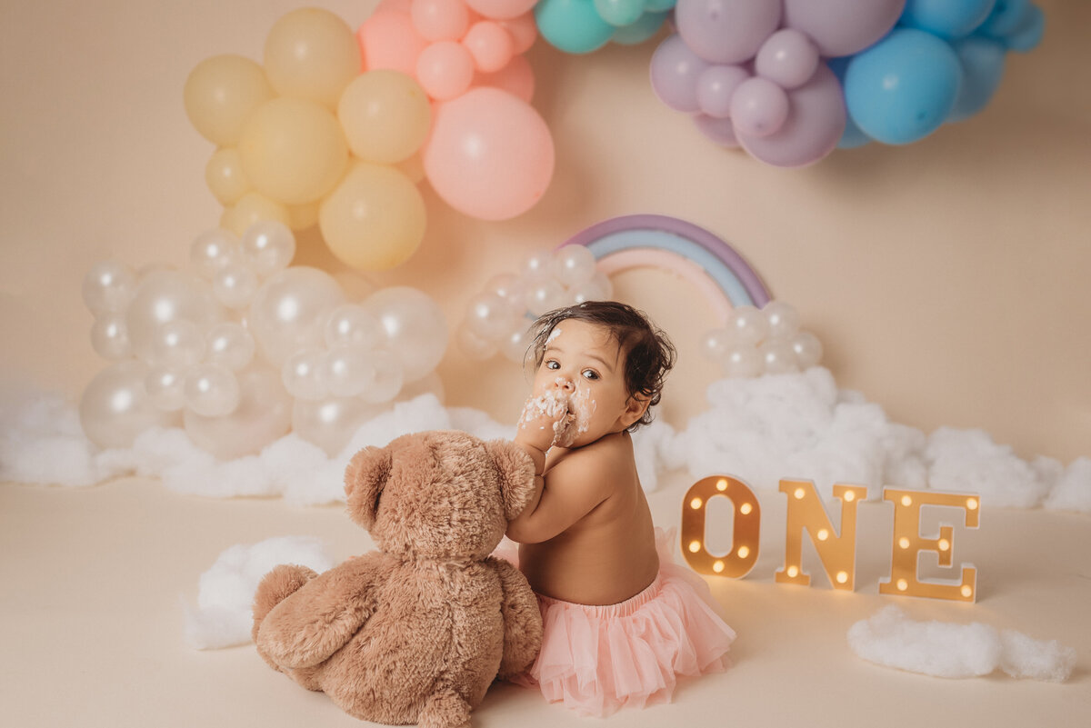 one year old baby girl sitting on tan backdrop wearing pink tutu eating cake and looking over shoulder with teddy bear sitting next to her. Rainbow pastel balloons in background.