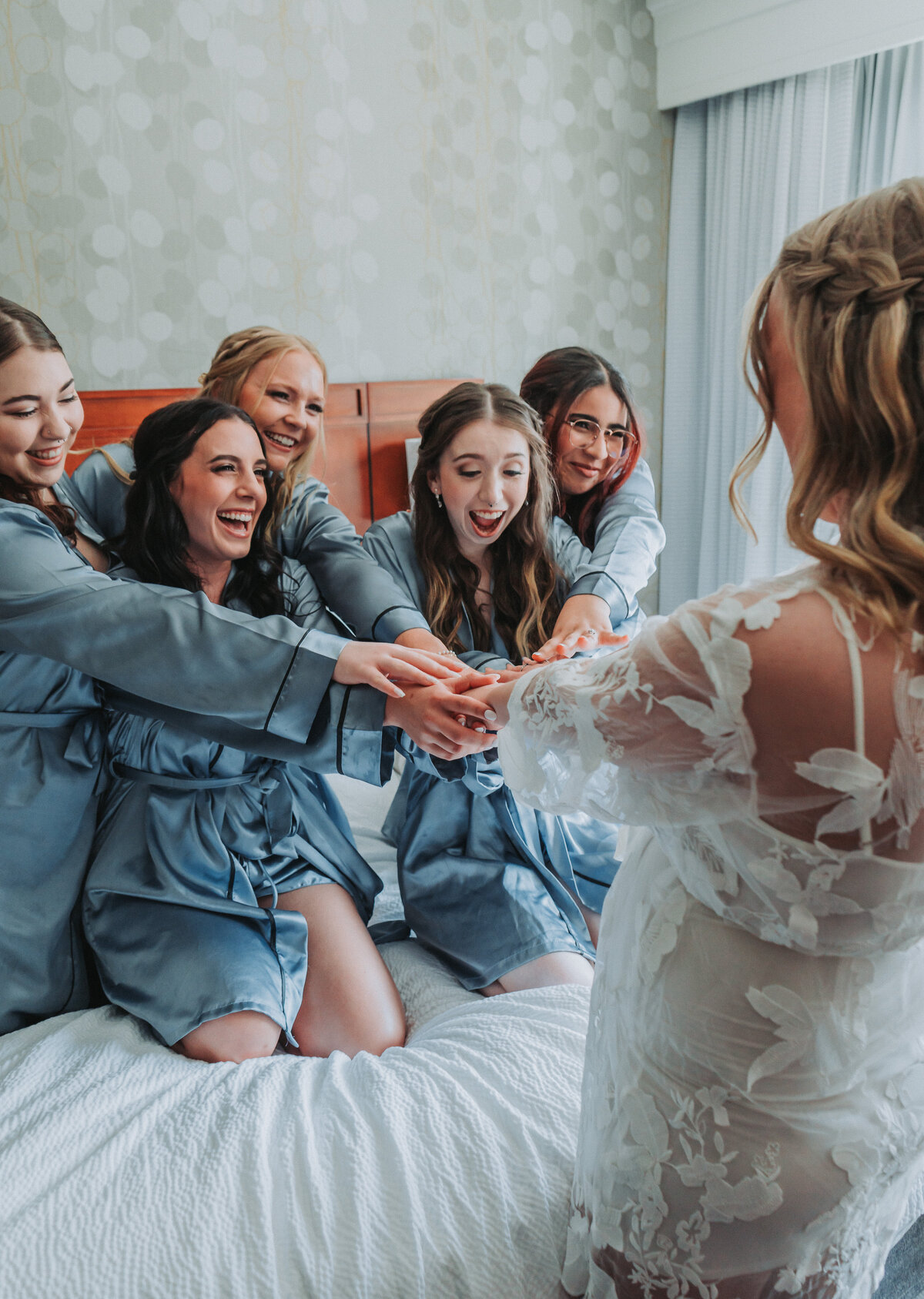 A joyful bride and her bridesmaids in matching blue robes are cheering and laughing together on a bed during wedding day preparations at the Hampton Roads Event Center in Newport News, Virginia, captured by Quinn Photography LLC.