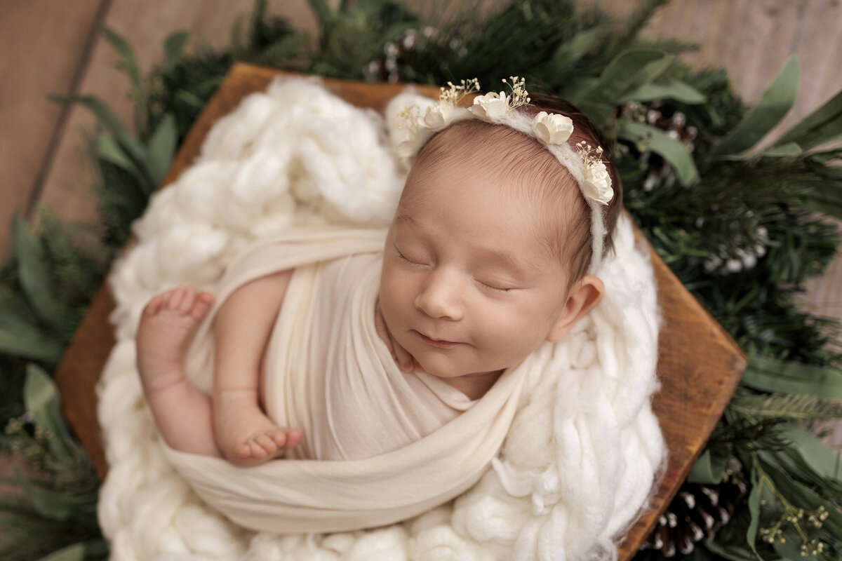 Newborn baby swaddled in a cream blanket, smiling slightly, and lying in a wooden bowl surrounded by greenery and pinecones, wearing a floral headband.