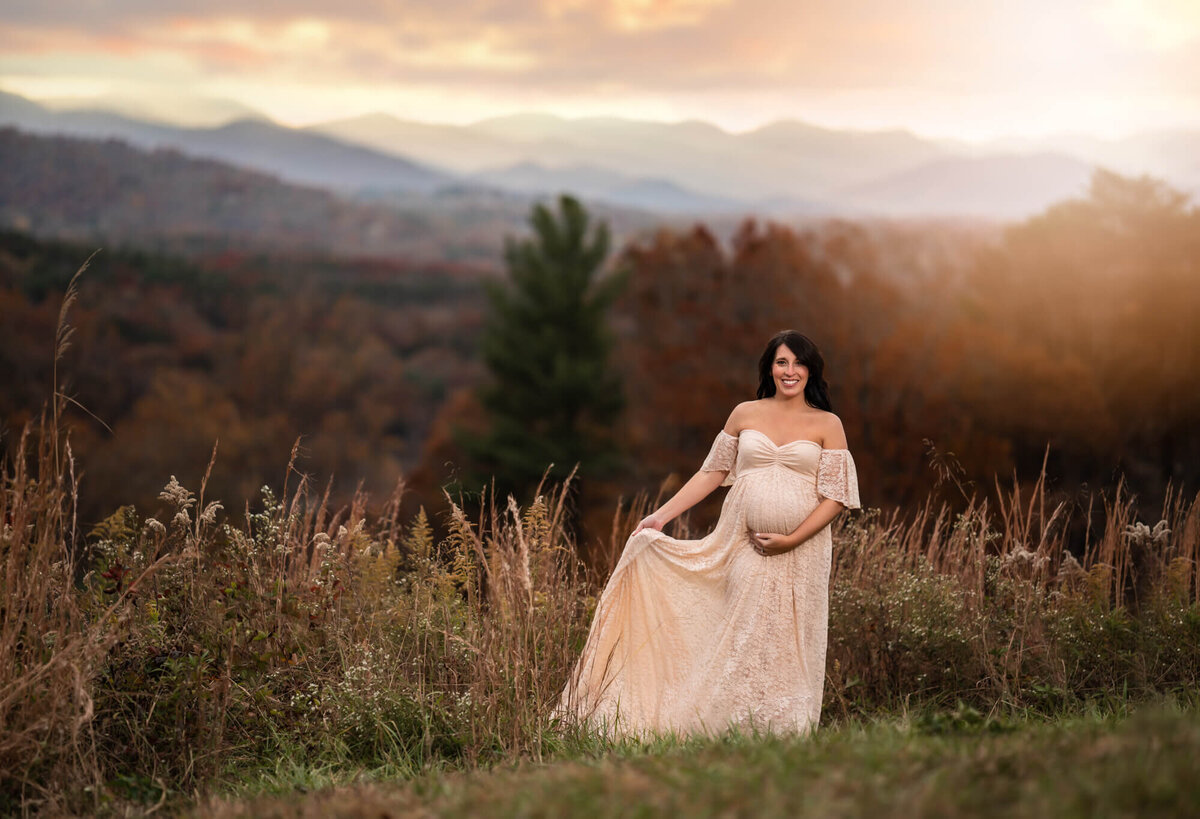 An expecting mama to be  swishes her long white dress and laughs while standing in a field with mountain in the distance near Asheville, nC