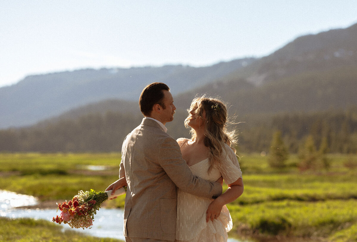 bride holds flowers and hugs the groom as they stand in front of the creek in a green, glowing meadow