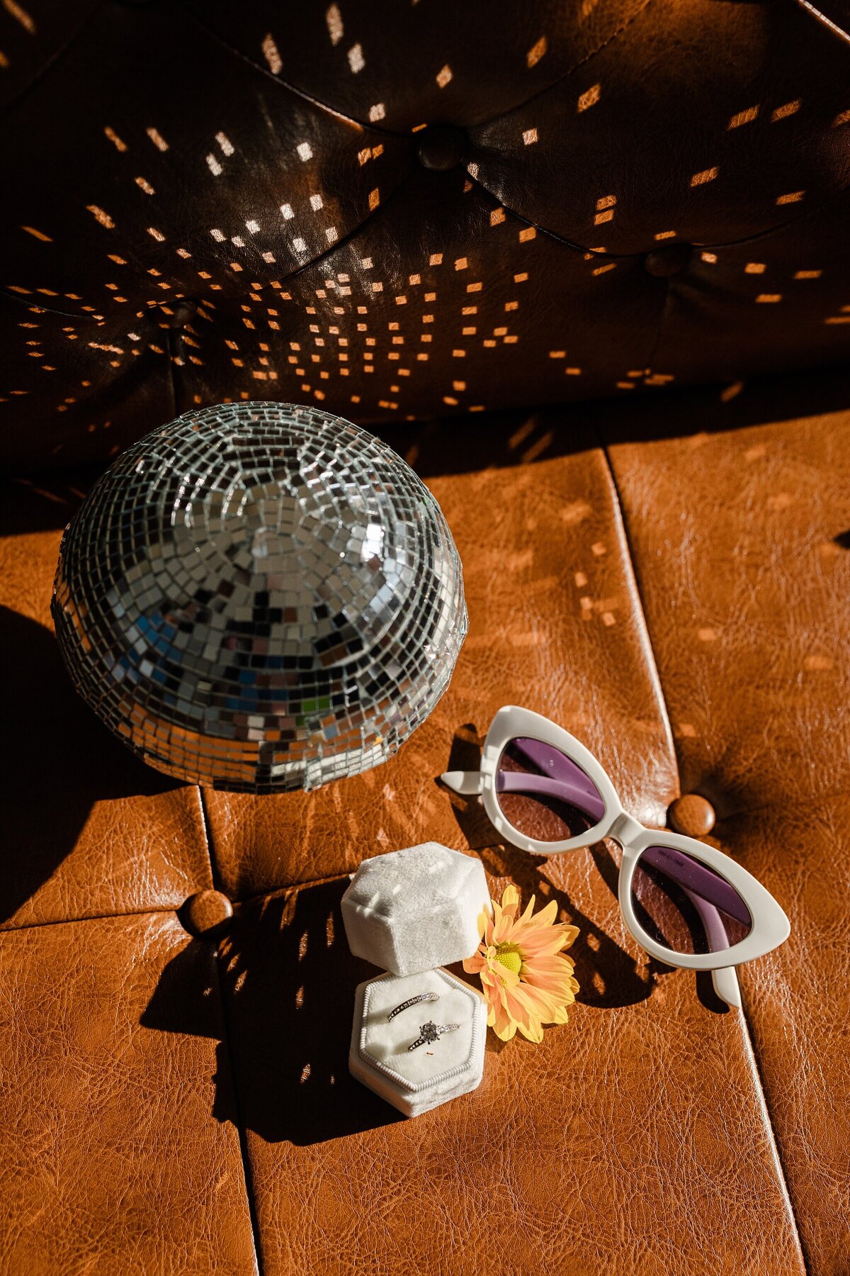 Detail shot of two rings in a white hexagonal ring box, a yellow flower, trendy white and purple sunglasses, and  disco ball resting on a leather couch. Light from the disco ball is reflecting interesting patterns onto the background.