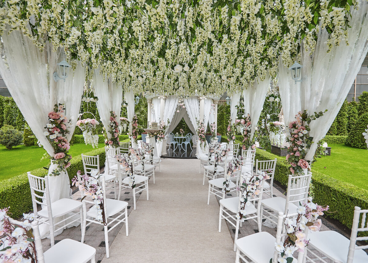 A luxurious wedding ceremony is set with a floral ceiling of wisteria with white drapes and chairs.