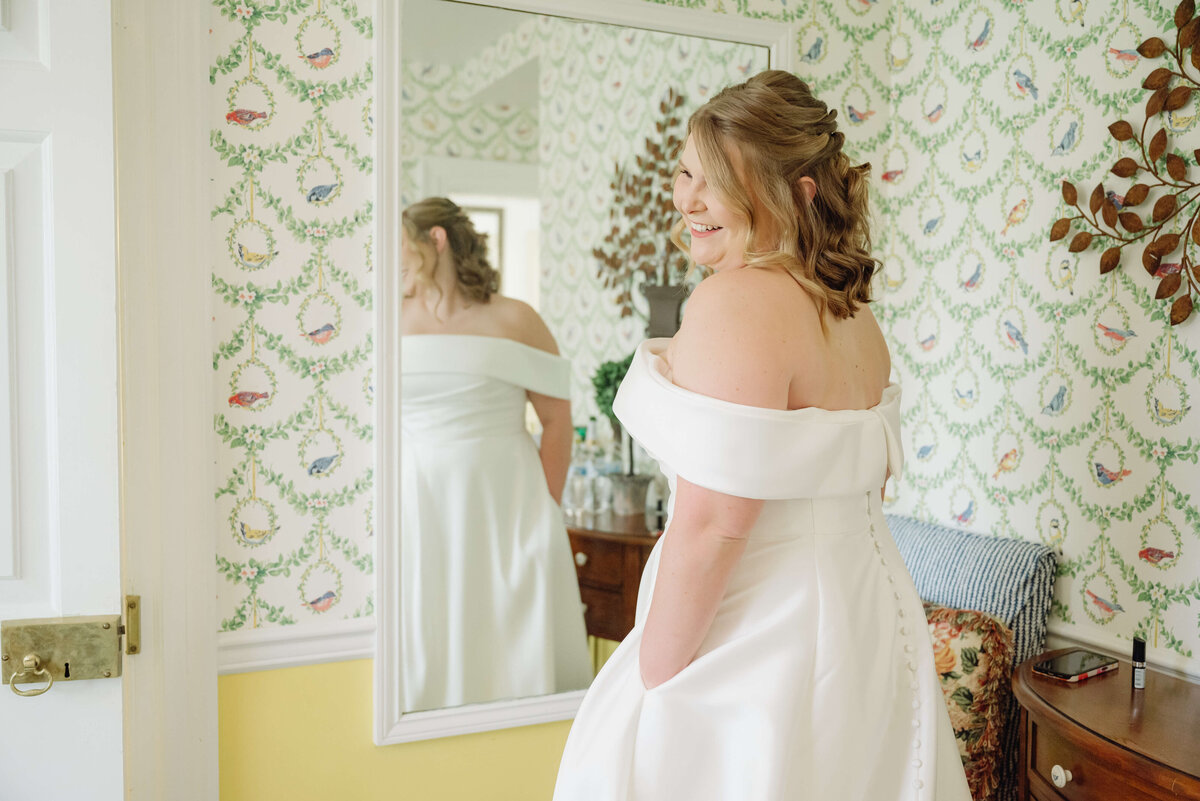 bride in a luxury off the shoulder wedding dress standing in front of a mirror in her bridal suite at middleburg community center and laughing over her shoulder
