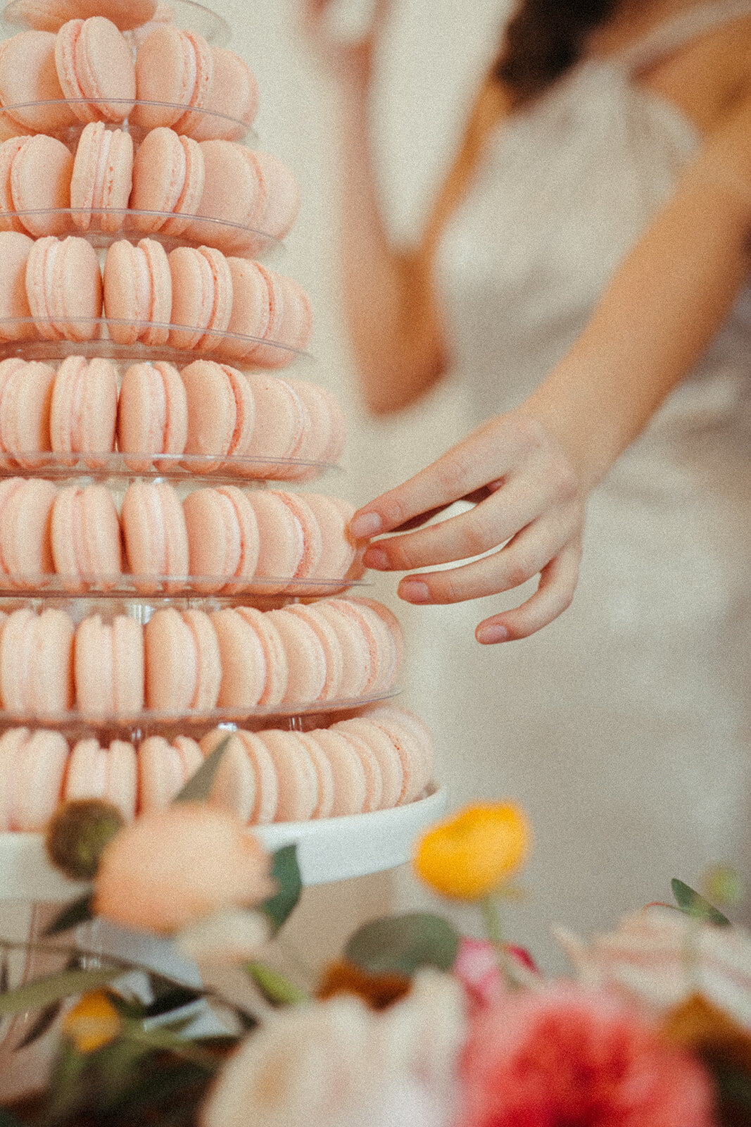 A close-up of the bride reaching into a tower of peach-colored macaroons setting on a cake stand next to flower arrangement.