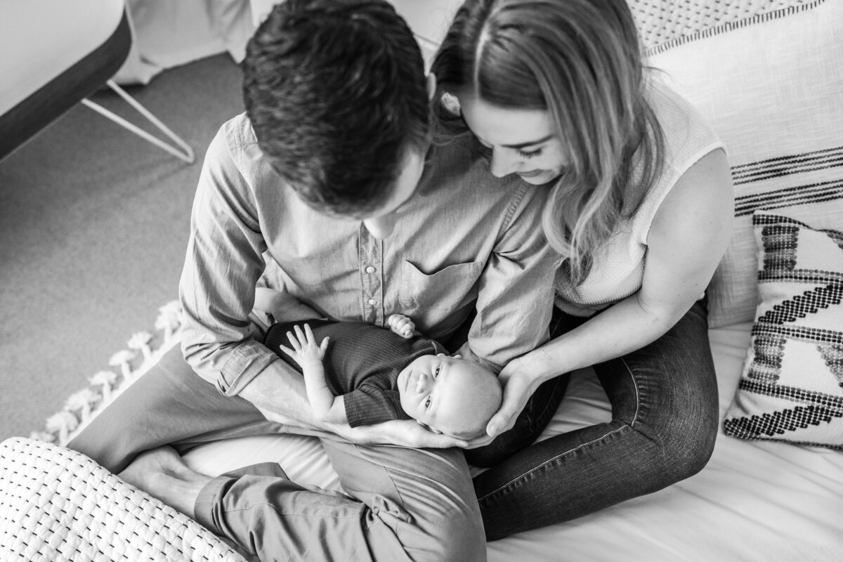 A joyful toddler dances on a bed while his parents, sitting nearby with a newborn in their arms, smile and watch.