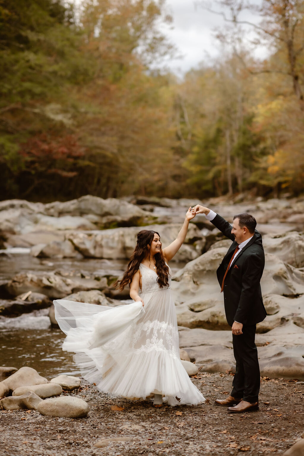 Bride twirling in a flowing lace wedding dress as the groom holds her hand by the river at Greenbrier, capturing a joyful moment during their eloping to Gatlinburg celebration surrounded by autumn forest.
