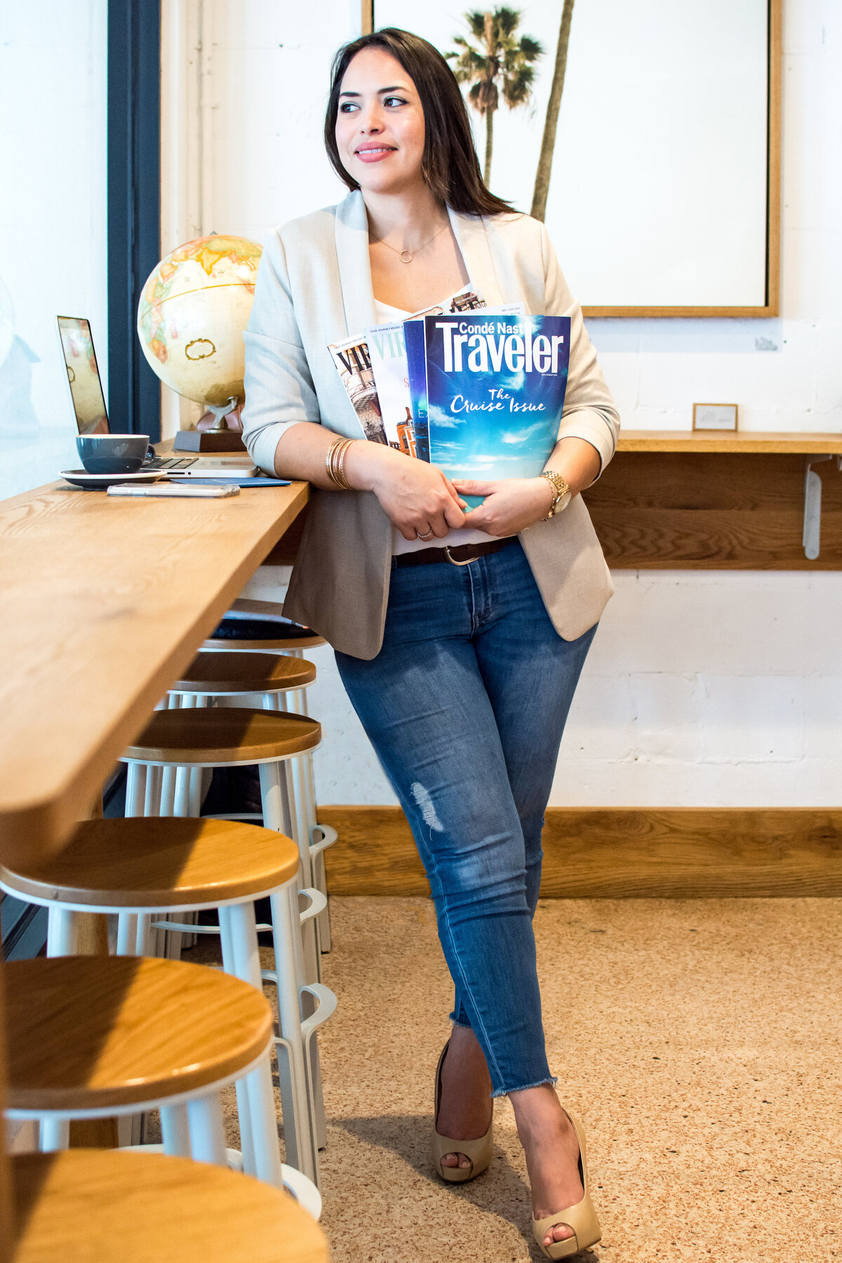 Hispanic woman wearing tan blazer and jeans leans agains a coffee shop bar holding travel magazines