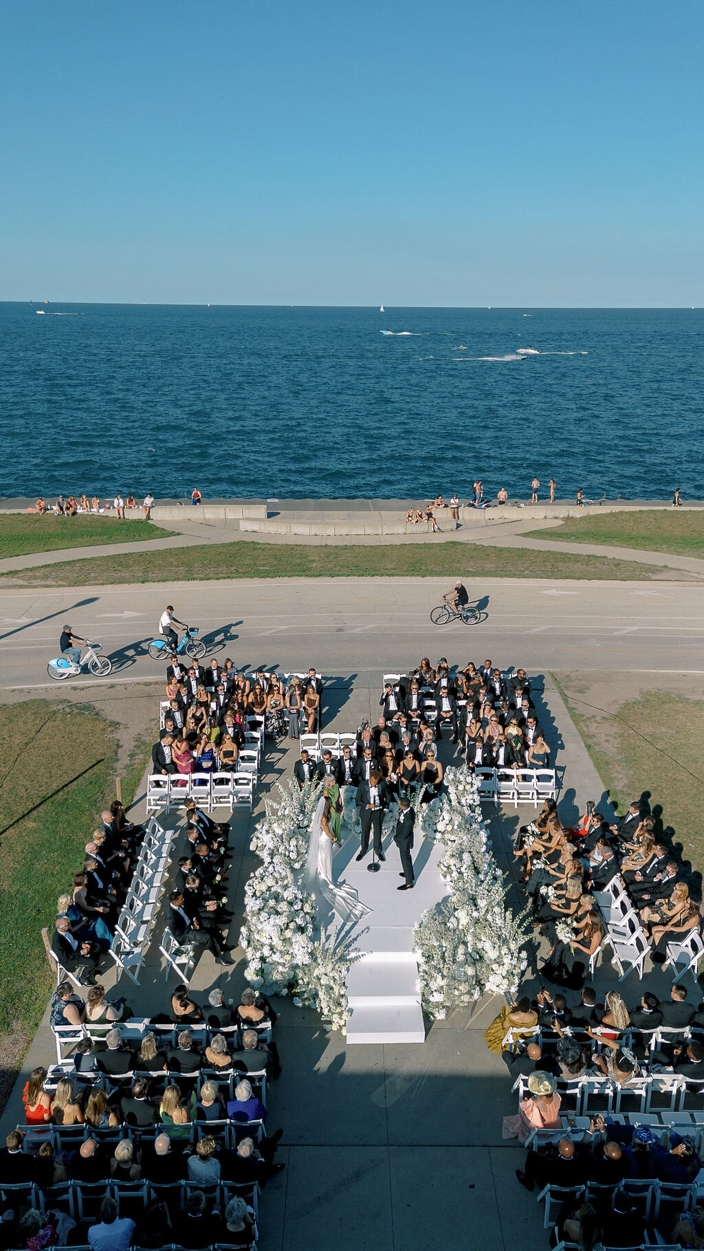 Outdoor wedding ceremony on Chicago's Lakefront Trail with Lake Michigan in the background. Guests are seated in formal attire while cyclists pass by on the adjacent path.