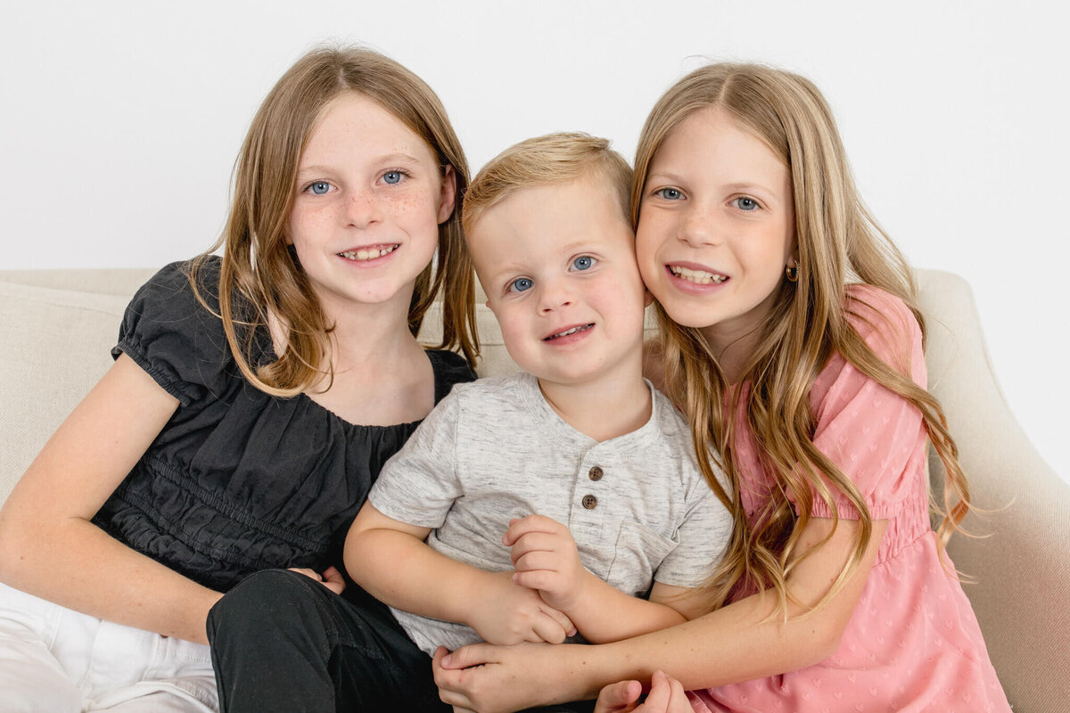 Two blonde sisters hugging their little brother, smiling at the camera against a white background.