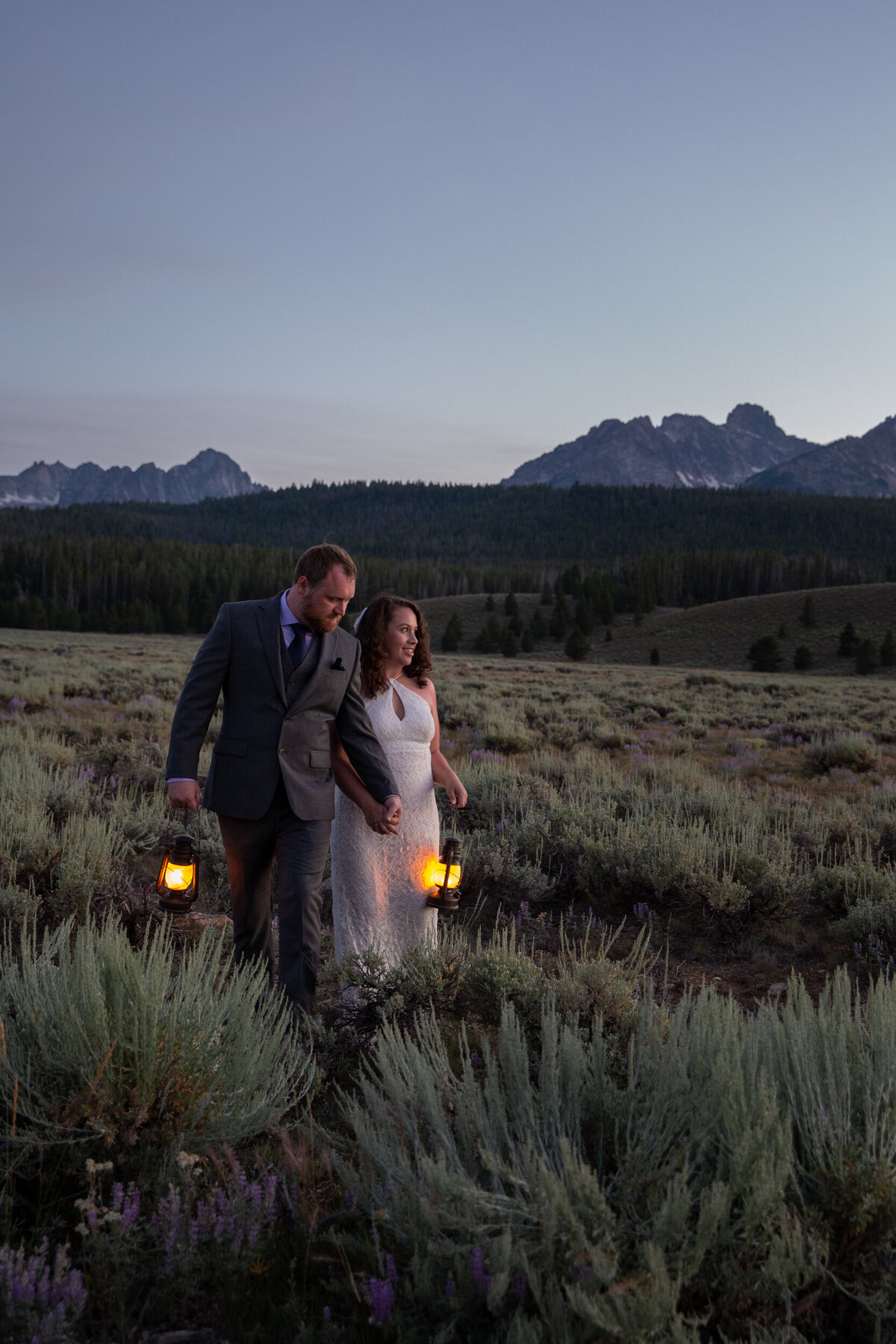 A bride and groom walk through a grassy meadow with lanterns in their hands as the sunsets.