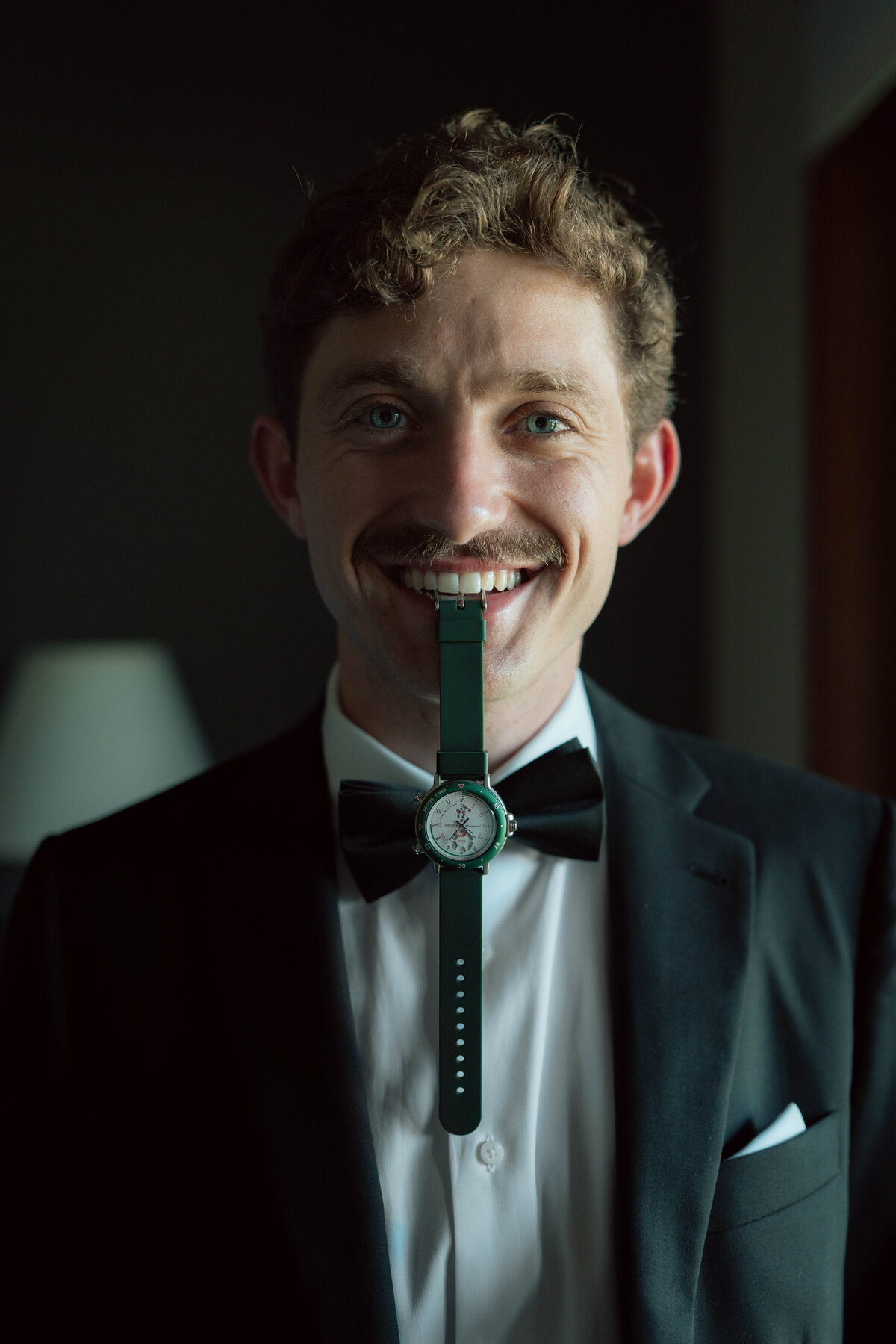 Groom portrait in tuxedo with green wristwatch held in mouth