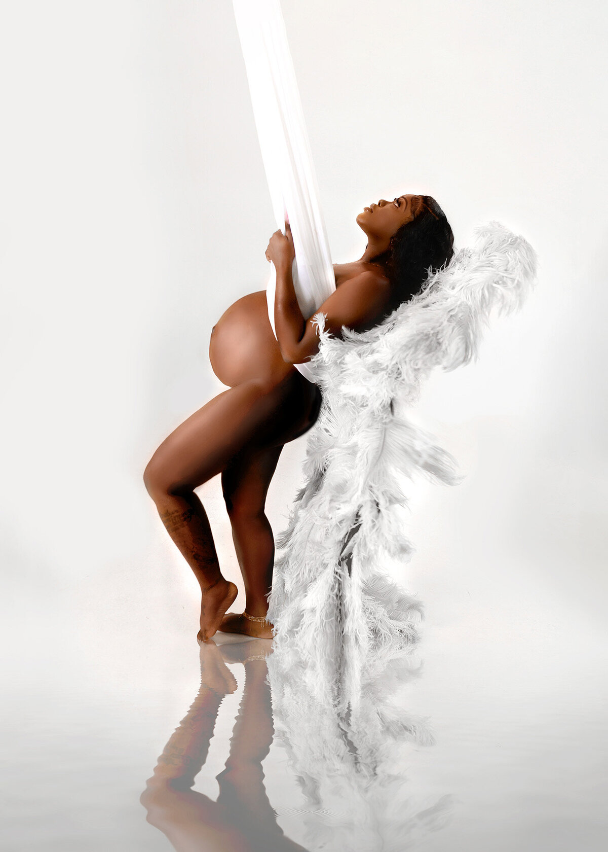 Pregnant woman posing nude leaned back in a silk fabric suspended from the ceiling wearing angel wings in all white studio set up
