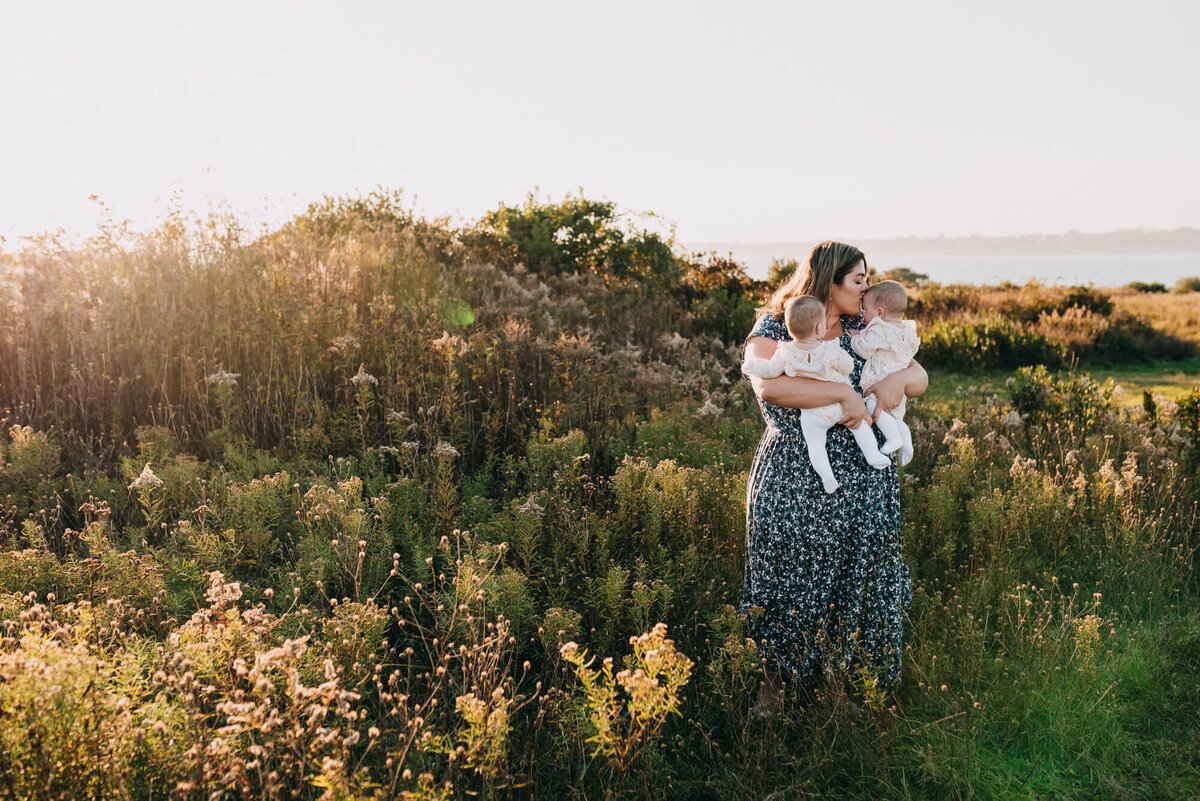 A mother wearing a blue and white floral dress holds her twin daughters in a field at sunset during their photoshoot with Allison Wolf, Boston Family Photographer