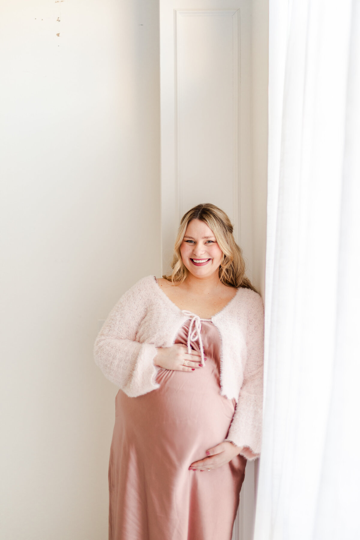 A radiant mother in an elegant silk dress, cradling her baby bump while leaning against a sunlit window, smiling with anticipation.