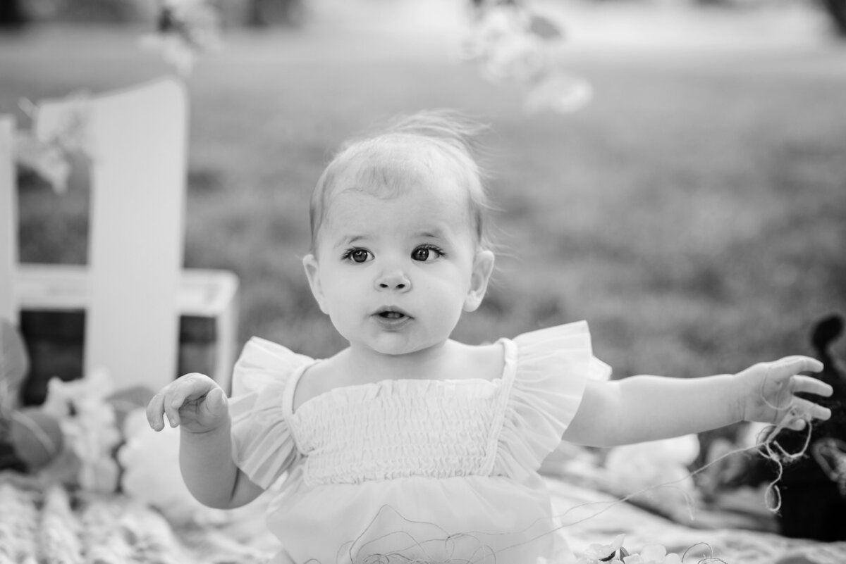 black and white image of wide eyed one year old baby girl sitting outdoors in front of  a number one sign and florals