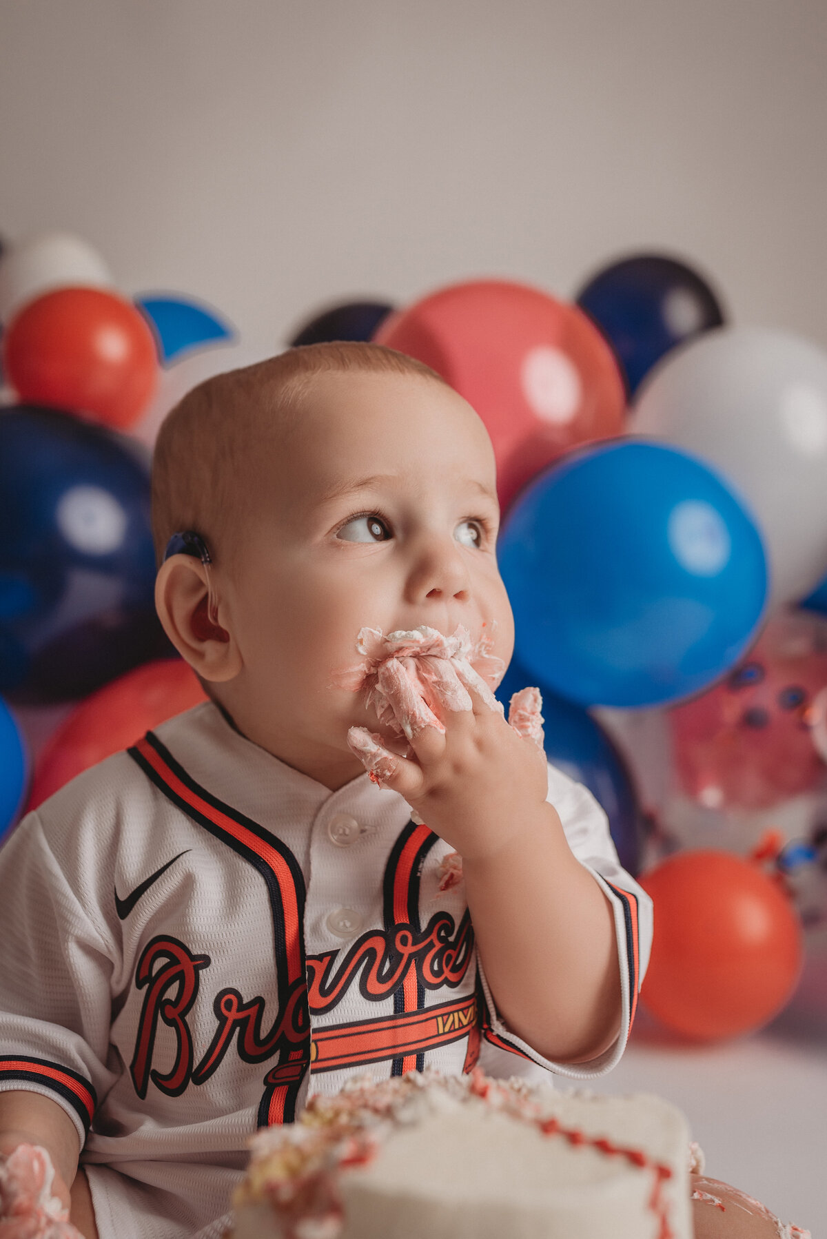 One year old baby boy sitting on floor eating birthday cake with Atlanta Braves baseball jersey on and red, white, blue balloon garland behind him