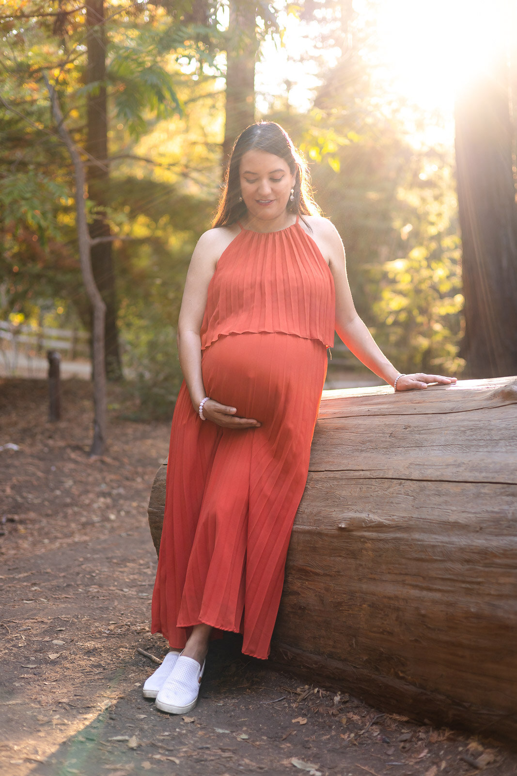 Full-body portrait of a pregnant woman leaning on a log with sun flare behind her – Ellobelle Photography