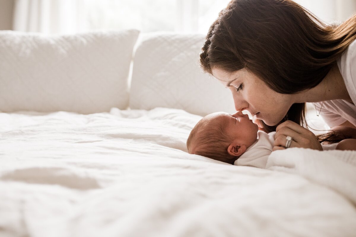 A mother affectionately touching noses with her newborn baby on a white bed during an at-home newborn photography session.