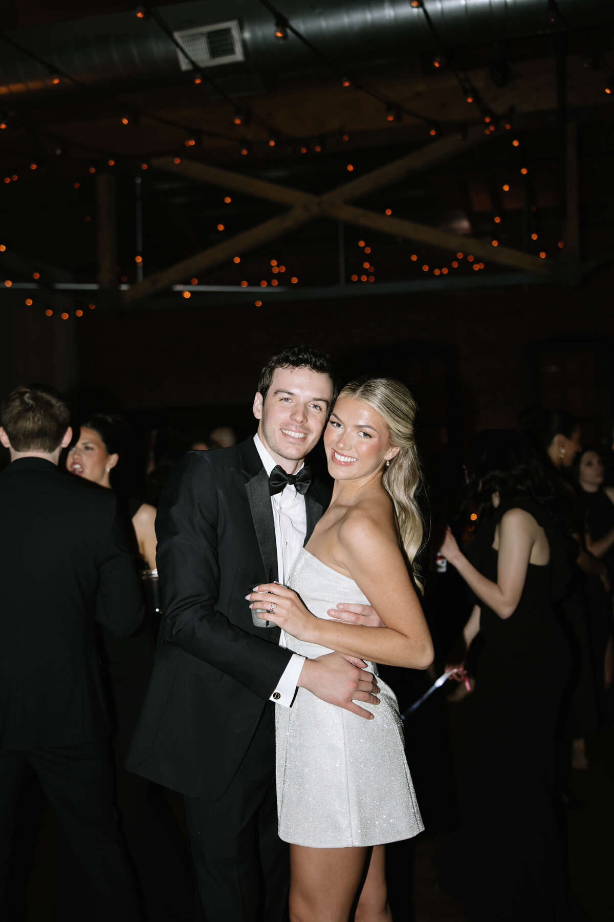 A groom in a beige suit kisses the bride's hand as she smiles, holding a white parasol. They're on a sunny palm-lined path with tropical foliage.