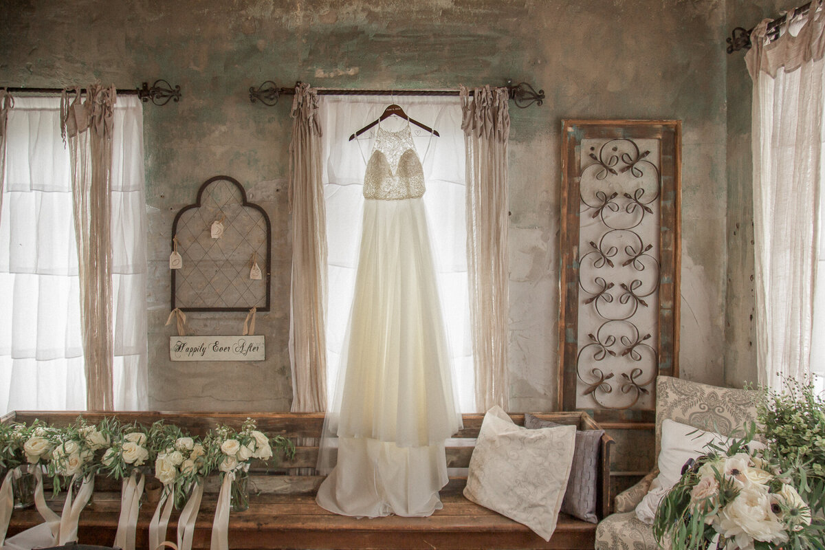 A wedding dress hangs in a rustic room with white curtains, surrounded by floral bouquets and vintage decor.