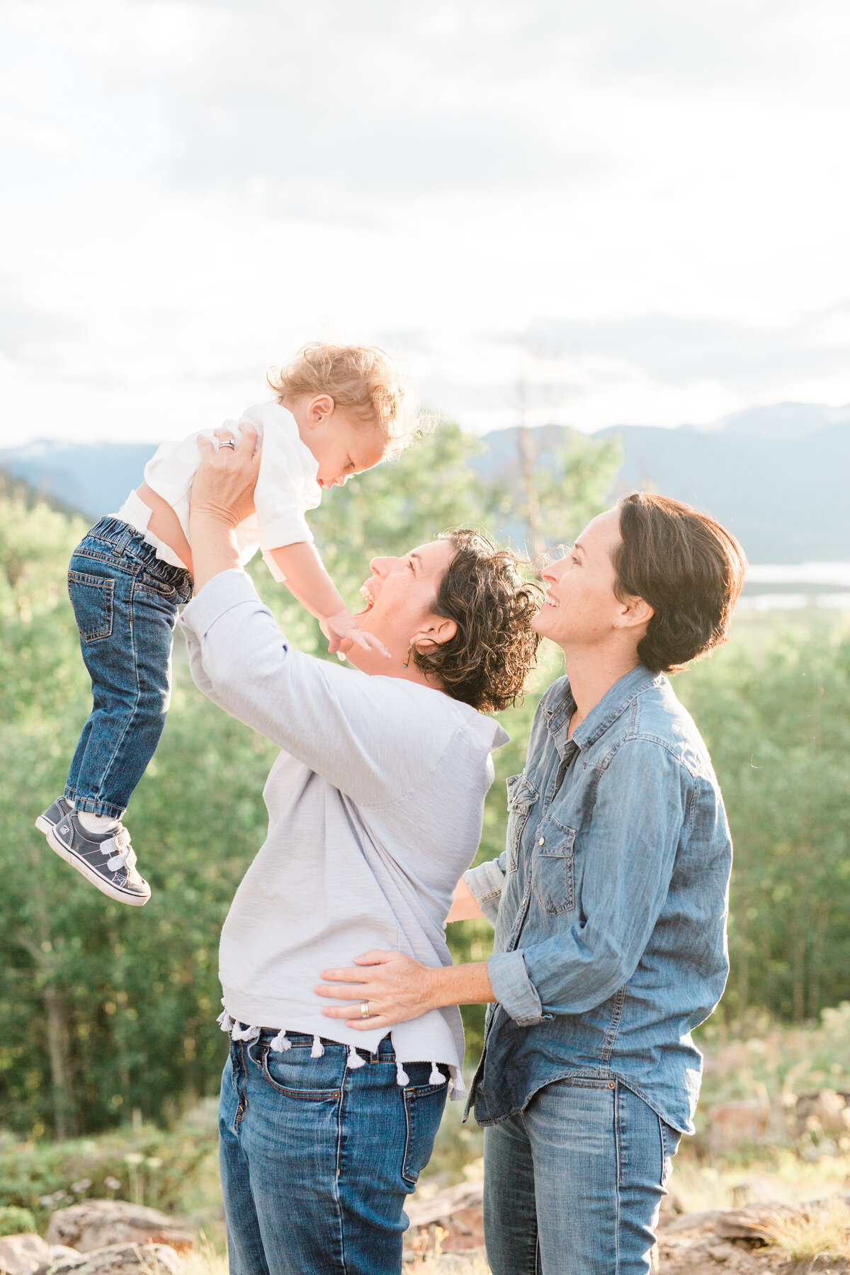 Two moms hold their son together above their heads. They are all smiling with an overlook of mountains behind them