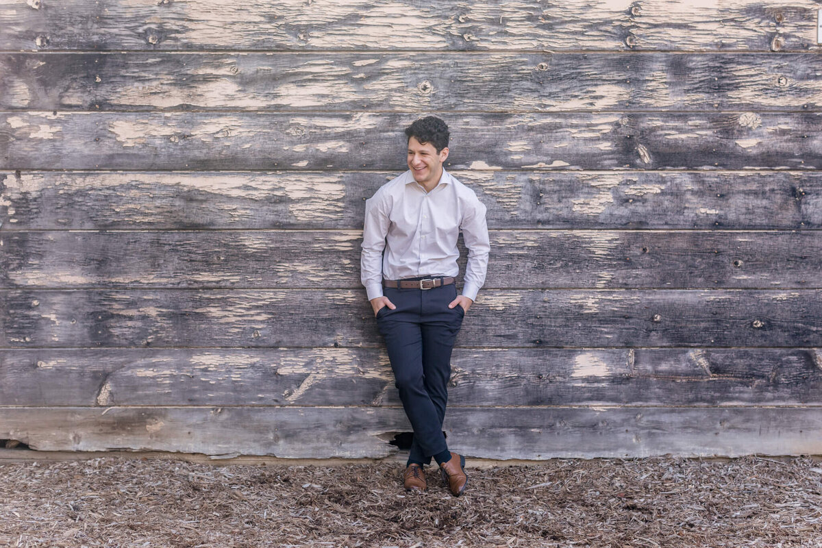 A young man with brown hair leaning against a wooden wall, standing with his hands in the pockets of his navy dress pants and looking into the distance smiling.