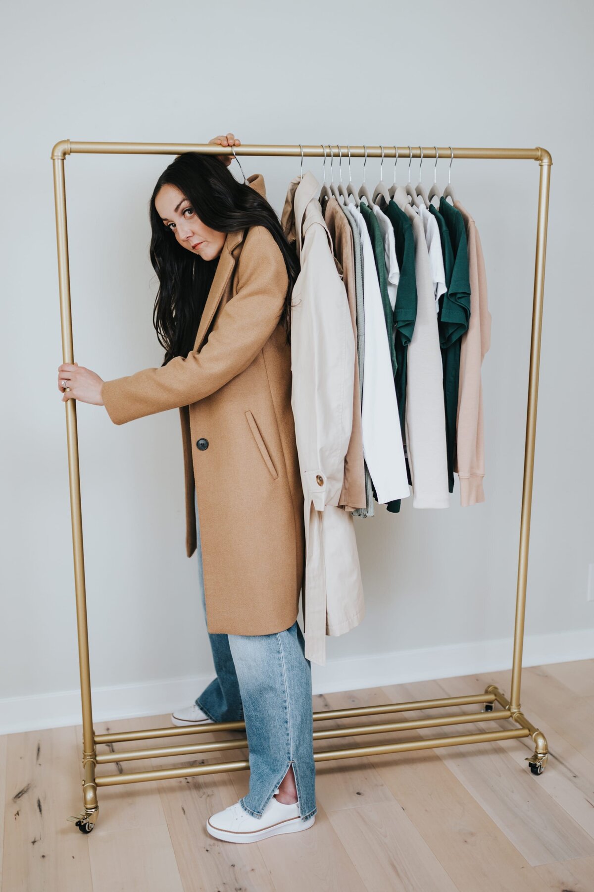 Woman with long, brown hair wears a camel-colored trenchcoat that hangs from a hanger on a clothing rack
