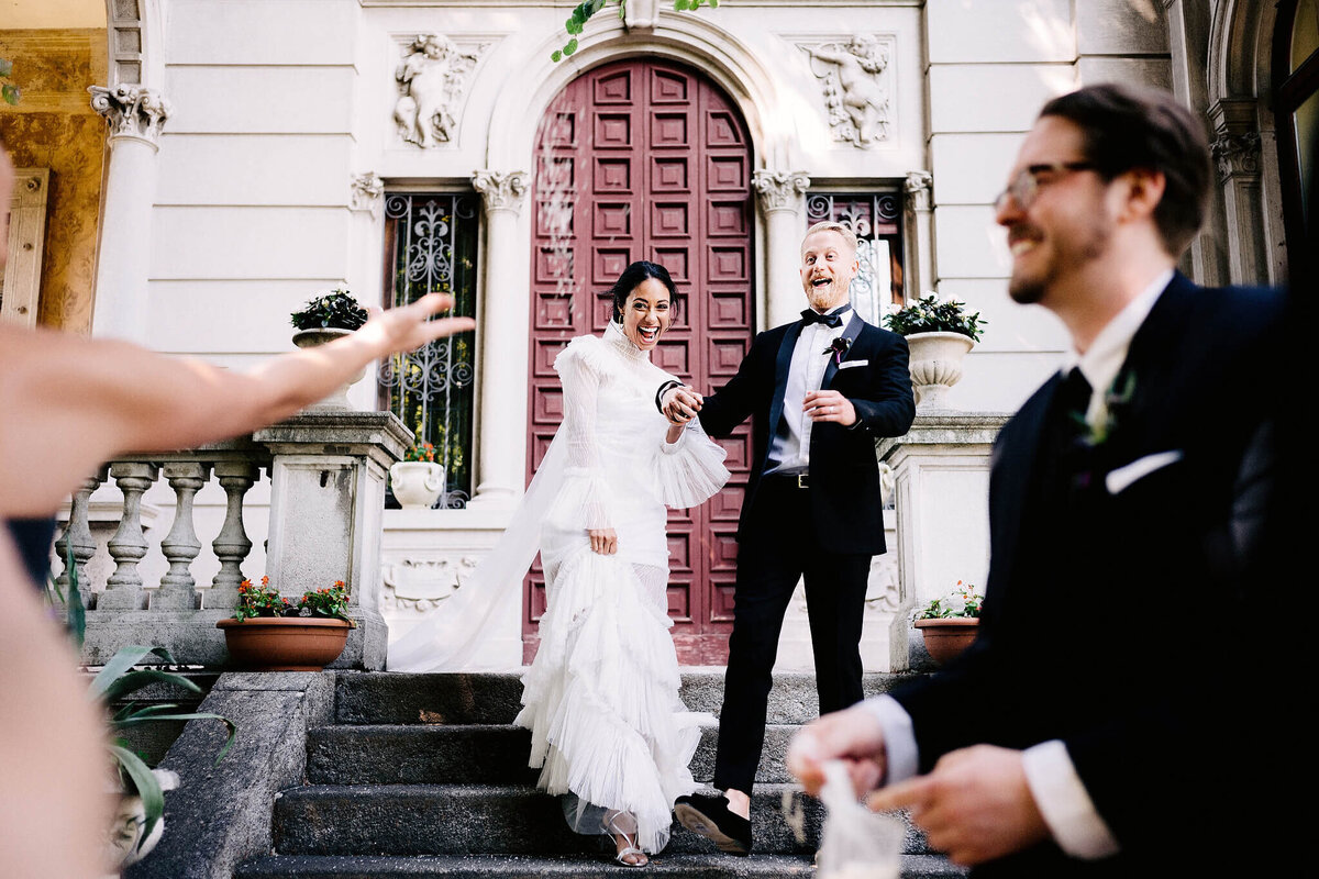 Bride and groom, holding hands happily while going down the staircase of a vintage villa, as guests throw confetti at them