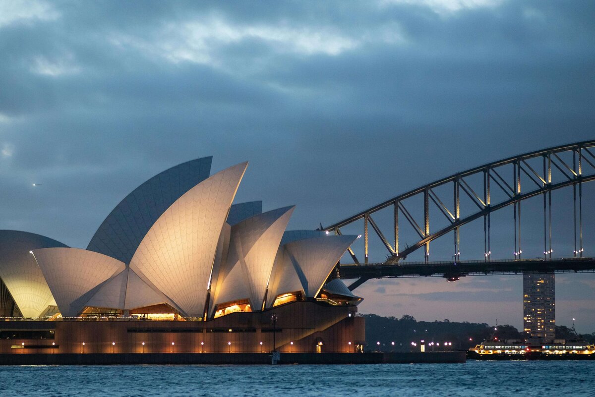 A breathtaking view of the Sydney Opera House at sunset, with its white sails glowing against the orange and golden hues of the sky. The serene water and intricate railing add to the beauty of this unforgettable destination, showcasing the global adventures made possible through vacation fundraising for charities.