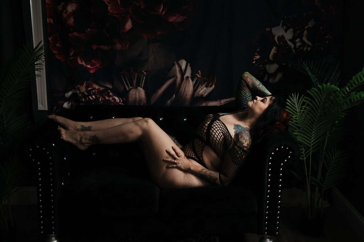 A woman in black mesh lingerie lays across a black couch under a floral tapestry