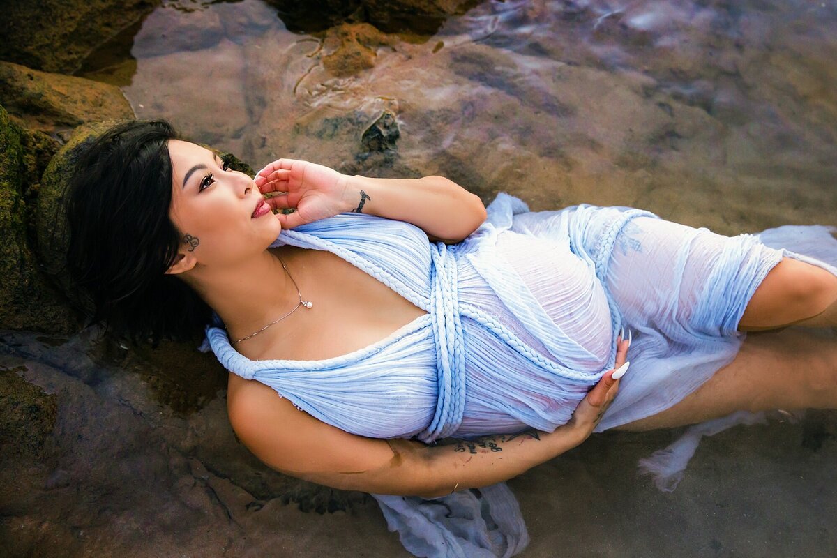 Beautiful woman with black hair and blue maternity gown lays in rock pool in Makena during her Hawaii babymoon portrait session