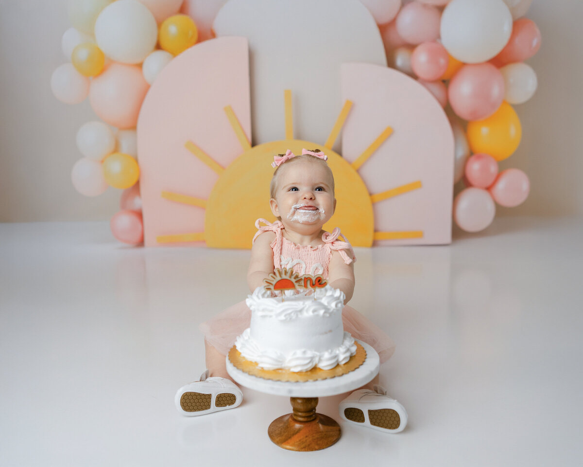 Baby in peach “one” dress smiling with cake during sunrise-themed cake smash. Photograph by Yucaipa cake smash photographer Kaitlyn Dawn Photography.