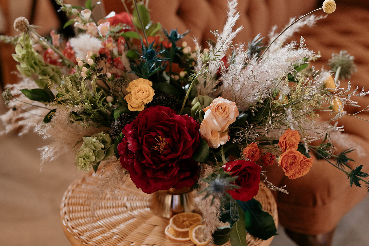 A richly textured floral arrangement with deep red and peach blooms, greenery, and dried elements set on a woven table, blending rustic and romantic aesthetics.