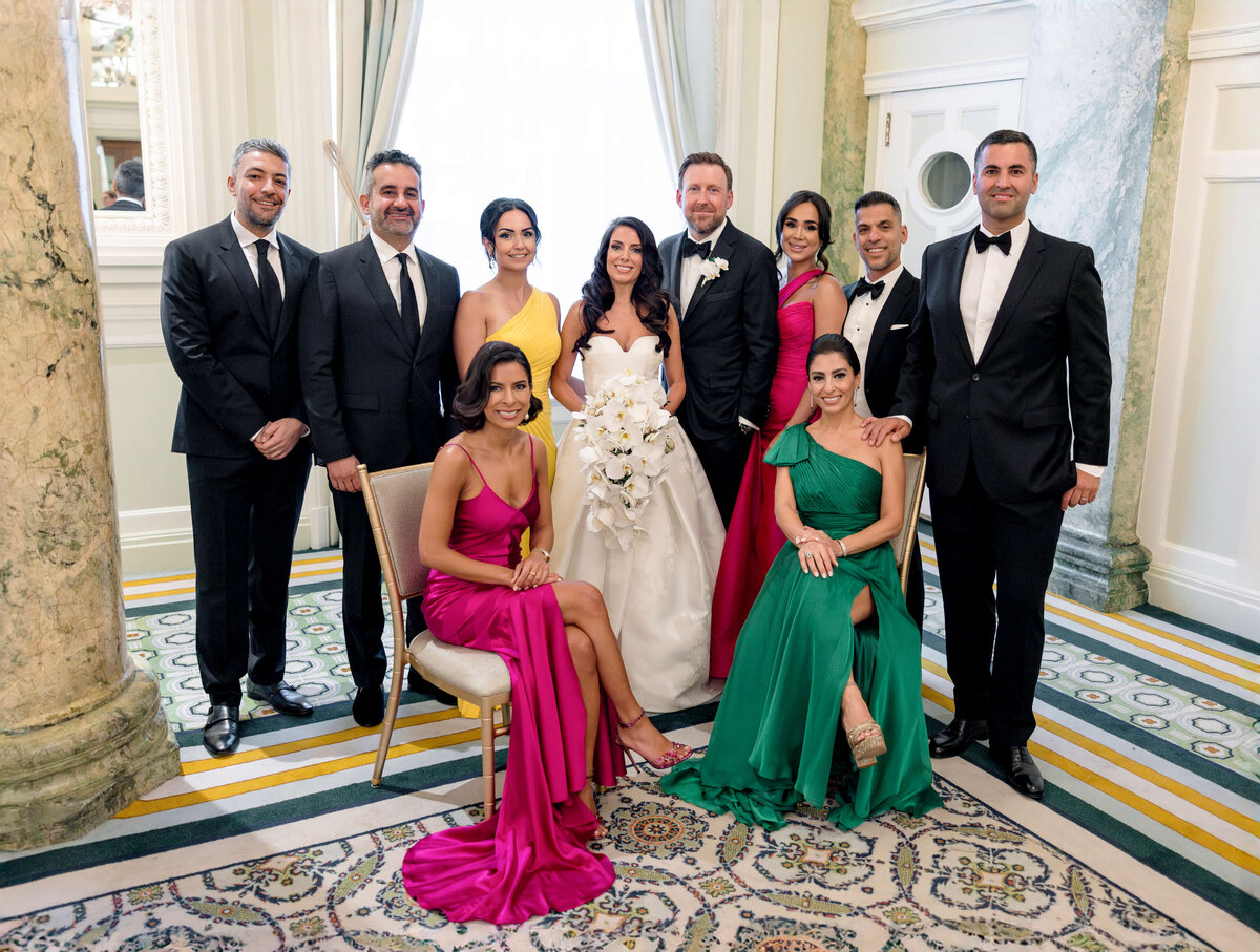 A group photo of a wedding party with seven people standing and three people sitting in a formal room. The bride is in the center wearing a white gown, surrounded by bridesmaids and groomsmen in elegant dresses and suits.